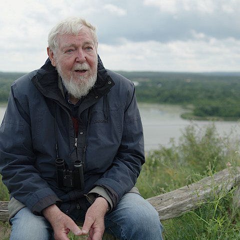 Prof. Michael Succow sitzt auf einem Baumstamm. Im Hintergrund ist ein See und Wald zu erkennen. 