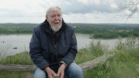 Prof. Michael Succow sitzt auf einem Baumstamm. Im Hintergrund ist ein See und Wald zu erkennen. 